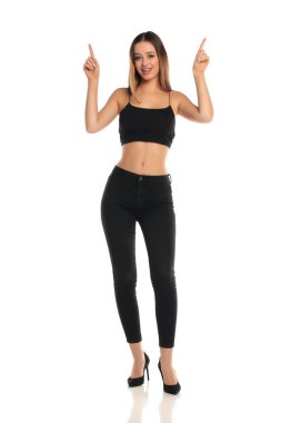 Uzun boylu esmer bir kadının uzun saçlı, siyah bir bluz, dar bir kot pantolon ve yüksek topuklu ayakkabıyla iki eliyle gülümseyen ve yukarı doğru bakan beyaz bir arka planda mesaj ya da ürün gösteriyormuş gibi..