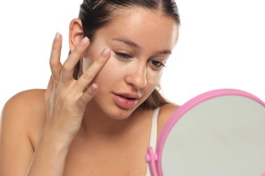 Genç bir kadın, aynaya bakarken gözlerinin altına kapatıcı sürüyor. Stüdyo resmi beyaz arka planda..