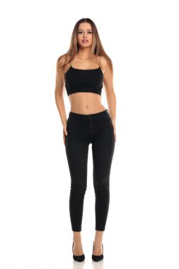 Uzun boylu, düz saçlı, siyah bluzlu, dar kotlu ve yüksek topuklu giyen uzun boylu esmer bir mankenin beyaz arka planda güvenle durduğu bir fotoğraf..