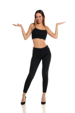 Uzun boylu esmer bir kadının uzun saçlı, siyah bir bluz, dar bir kot pantolon ve yüksek topuklu ayakkabı giymiş, gülümseyen ve avuçlarını havada tutan beyaz bir arka planda görünmez ürünler sunar gibi..