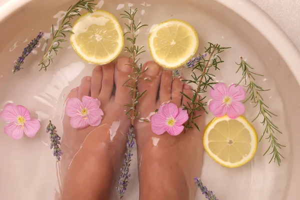 Doğal spa tedavisi için kadın ayaklarının limon dilimleri, biberiye, lavanta ve pembe çiçeklerle sırılsıklam olduğu bir stüdyo fotoğrafı.