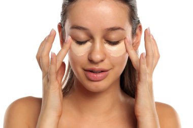 Gözlerinin altına kapatıcı süren genç bir kadın. İki eli kapalı. Stüdyo resmi beyaz arka planda..