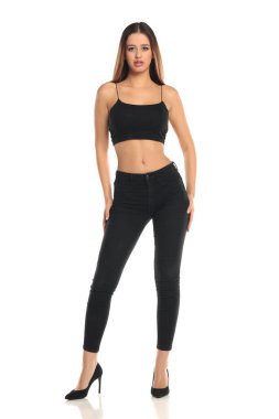 Uzun boylu, düz saçlı, siyah bluzlu, dar kotlu ve yüksek topuklu giyen uzun boylu esmer bir mankenin beyaz arka planda güvenle durduğu bir fotoğraf..