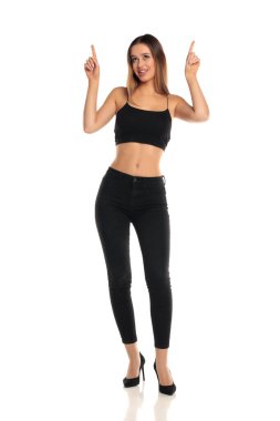 Uzun boylu esmer bir kadının uzun saçlı, siyah bir bluz, dar bir kot pantolon ve yüksek topuklu ayakkabıyla iki eliyle gülümseyen ve yukarı doğru bakan beyaz bir arka planda mesaj ya da ürün gösteriyormuş gibi..