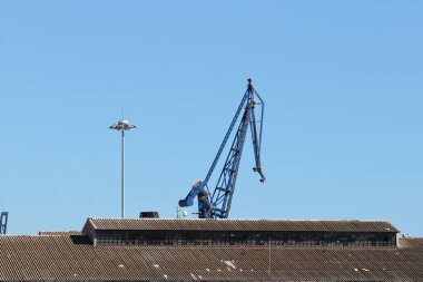 Endüstriyel liman vinci açık mavi gökyüzüne, kentsel liman altyapısına ve şehir parçasına karşı depo çatısından yükseliyor..