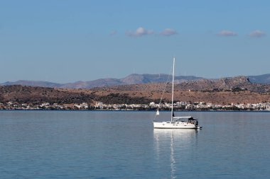 Sakin denizde yüzen yelkenli, kıyı kasabası ve arka planda tepeler, huzurlu yaz seyahatleri ve deniz hayatı..