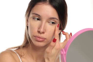 Beyaz bir arka planda, aynaya, portreye, vesikalığa bakarken gözünün altındaki kapatıcıyı parmaklarıyla harmanlayan esmer bir kadının stüdyo fotoğrafı..