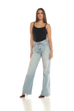 Siyah kolsuz tişörtlü, geniş paçalı, yüksek topuklu, kendinden emin beyaz arka planda duran genç bir kadının stüdyo çekimi..