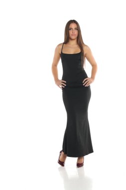 Stüdyo resmi, siyah saçlı, uzun siyah elbiseli, yüksek topuklu giyen, elleri kalçasında, tüm vücudunda, beyaz bir arka planda duran bir kadın.