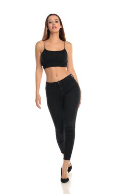 Uzun boylu, esmer, uzun saçlı, siyah bluzlu, dar kotlu ve yüksek topuklu bir kadının beyaz arka planda güvenle yürüdüğü bir stüdyo resmi..