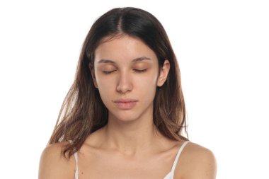 Beyaz bluzlu, makyajsız, gözleri kapalı, beyaz arkaplanda portresi olan esmer bir kadının stüdyo fotoğrafı..