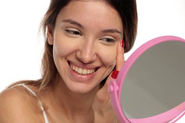 Gülümseyen esmer bir kadının, beyaz bir arka planda, aynaya, portreye, vesikalığa bakarken gözünün altındaki makyajı parmaklarıyla harmanladığı bir resim..