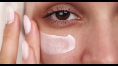 4k video, yavaş çekim, bir kadının yüzünün stüdyo çekimi gözlerinin altına cilt kremi sürerken, hijyen ve kozmetik konsepti hafif arka planda.