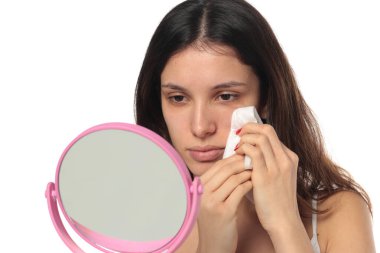 Beyaz arka plandaki aynaya bakarken yüzünü ıslak mendille temizleyen bir kadının stüdyo fotoğrafı..