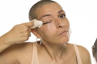 Kolsuz tişörtlü, kel bir kadının beyaz arka planda dokusu olan göz makyajını temizlerken çekilmiş bir fotoğraf.