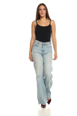 Stüdyo resmi, kahverengi saçlı, siyah kolsuz bluzlu, açık mavi kotlu, yüksek topuklu, beyaz bir arka planda yürüyen bir kadın..