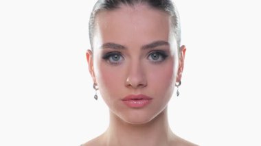 Ciddi bir genç kadın kameraya bakıyor ve iki kez göz kırpıyor. Güzellik portresi, beyaz arka planda stüdyo çekimi.