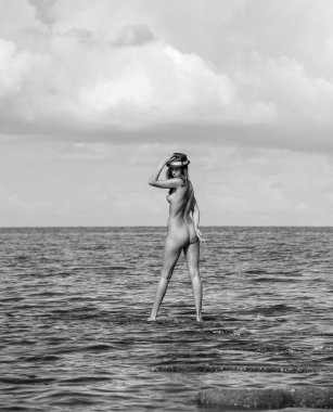 Siyah şapkalı güzel, çıplak bir kadın sahilde yaz tatilinin tadını çıkarıyor. Denizde çıplak poz veren seksi.