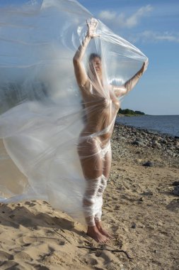 Genç çıplak kadın sahilde polietilen filmde poz veriyor.