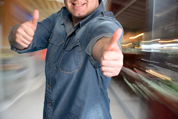 Young man gesturing thumbs up at shopping mall — Stock Photo, Image
