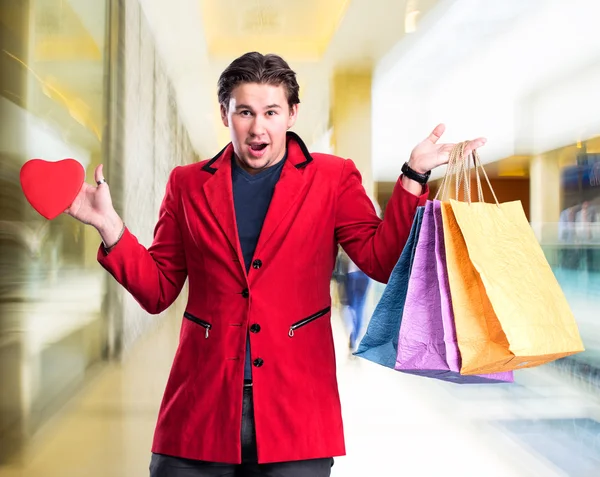 Sonriente hombre guapo sosteniendo bolsas de compras y corazón rojo —  Fotos de Stock