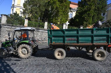 UZHOROD, UKRAINE - 19 Eylül 2025: İşçiler Uzhhorod 'da gündüz vakti kaldırımlı bir caddede traktör ve römorkla atık taşıyorlar