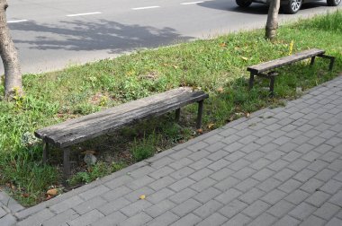 Bir yolun yanındaki kaldırıma iki ahşap bank konur. Çimenler ve küçük bitkiler sıraların etrafını sarar. Arabalar gün ışığında yolda gidiyor.