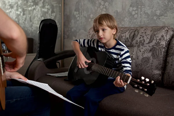Young male musician teaches little boy student how to play the acoustic guitar and play by notes