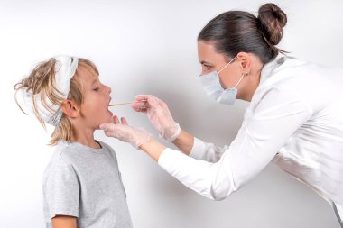 Tıp, sağlık ve pediatri konsepti. Tıbbi maskeli ve şeffaf eldivenli bir kadın doktor kafa yaralanması geçiren küçük hasta bir çocuğu muayene ediyor. Boğazını kontrol eder ve koronavirüs için örnek alır..