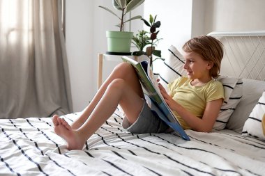 Sarı tişörtlü sarışın bir çocuk yatakta yatarken bir kitap okuyor.