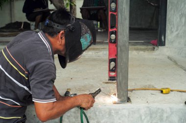 Yüz kalkanlı kaynakçı inşaat alanındaki beton zeminde çelik kolona kaynak yapıyor.