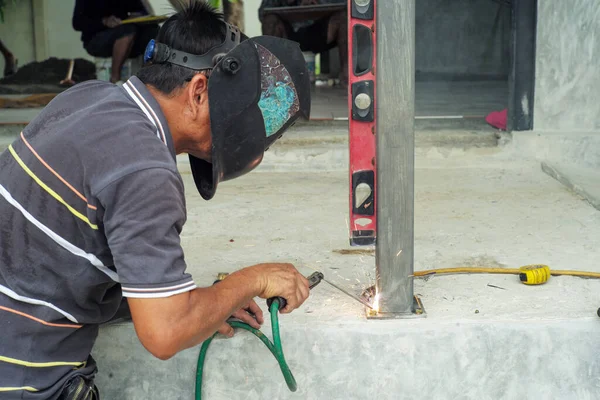 Welder with face shield is welding the steel column with the plate on the concrete floor at the construction site