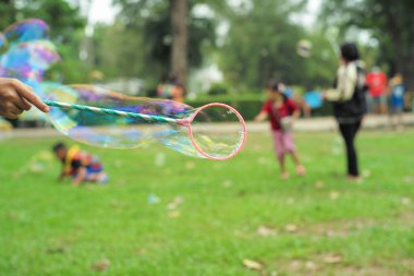 Parkta arka planda oynayan bulanık insan grubuyla sabundan baloncuk yapmak için plastik yüzüğü tutan el.