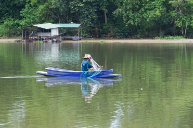 Yalnız adam teknede oturur ve arka planda ormanla birlikte ağdaki balıkları toplar..