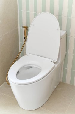 Açık kapaklı modern beyaz tuvalet ve temiz banyoda bide spreyi, dikey çizgili duvar fayansları, bej zemin ve doğal aydınlatma taze ve hijyenik atmosfer yaratır.