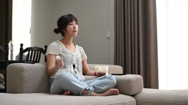 Mutlu Asyalı kadın evde huzurlu bir yaşam ve kişisel refahın tadını çıkarıyor. Oturma odasında yemek yerken rahat koltukta dinleniyor.