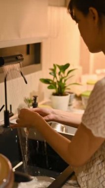 Basit ev yaşamını kucaklayan sakin Asyalı bir kadın, iyi bir hijyen için mutfak lavabosunda ellerini yıkarken rahatlatıcı ve kişisel bir rahatlama anı buluyor.