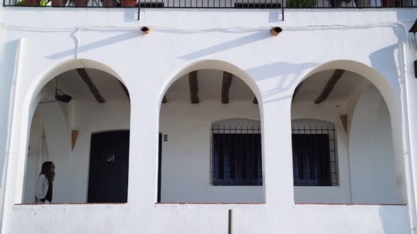 Jeune femme en manteau beige marchant sur la terrasse. Vue de l'arche à la fille aux cheveux longs. Journée ensoleillée sur la ville côtière. Village blanc dans une magnifique baie. Architecture méditerranéenne blanche. — Video