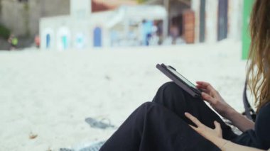 Siyahlı genç kadın sahilde bir e-kitap okuyor. Sayfaları değiştiren ellerin yakın çekimi. Denizde güneşli bir gün. Altın plajda sıcak tatiller. Yaz soğuğu. Beyaz kum ve eski tekne depoları. Tatil