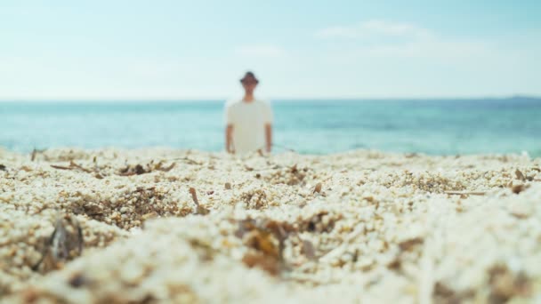Man is walking along the beach. Young man in a white T-shirt, black shorts and a black hat walks along the sand to the sea. Sunny summer day on the beach. Holidays in a warm country. Blue sea. — Stock Video