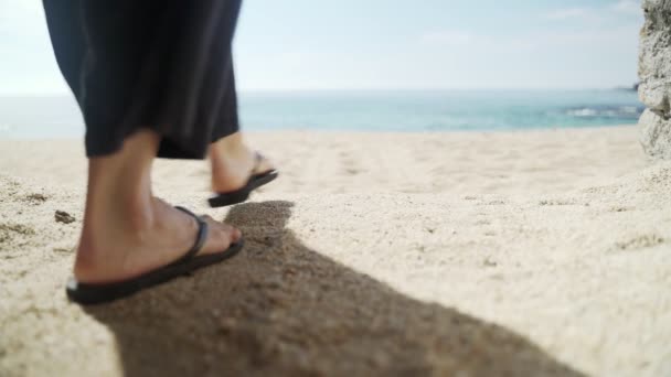 Mujer joven en pantalones negros anchos y un sombrero camina a lo largo de la playa hacia el agua. Día soleado en el mar. Primer plano de los pies en chanclas negras. Vacaciones cálidas en la playa dorada. Frío de verano. Arena blanca. — Vídeos de Stock