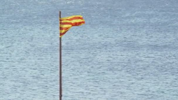 Vor dem Hintergrund des blauen Meeres flattert die Flagge Kataloniens im Wind. Wind an einem sonnigen Sommertag. Flagge mit gelben und roten Streifen. Leichte Wellen auf der Wasseroberfläche. Tourismus in Europa. — Stockvideo