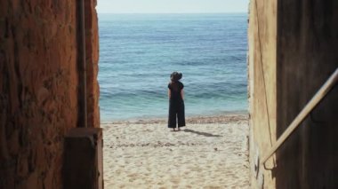 Geniş siyah pantolonlu ve şapkalı genç bir kadın sahile bakan eski bir şeritte duruyor ve deniz manzarasının tadını çıkarıyor. Kumsalda güneşli bir gün. Altın plajda sıcak tatiller. Yaz soğuğu. Beyaz kum.