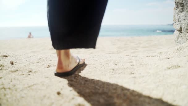 Geniş siyah pantolonlu ve şapkalı genç bir kadın sahile doğru yürüyor. Siyah parmak arası terliklerini çıkardı ve yalınayak gitti. Denizde güneşli bir gün. Ayaklara yakın çekim. Plajda tatiller. Yaz soğuğu — Stok video