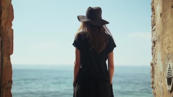 Geniş siyah pantolonlu ve şapkalı genç bir kadın sahile bakan eski bir şeritte duruyor ve deniz manzarasının tadını çıkarıyor. Kumsalda güneşli bir gün. Altın plajda sıcak tatiller. Yaz soğuğu. Beyaz kum. — Stok video