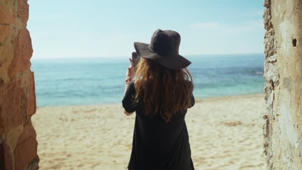 Ung kvinna i breda svarta byxor och en hatt står i en gammal fil med utsikt över stranden, har havsutsikt och göra foto med smartphone. Solig dag på stranden. Helgdagar. Sommarkyla. Vit sand. — Stockvideo