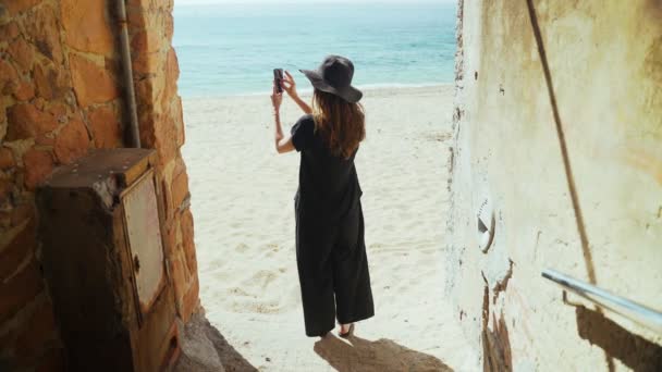 Ung kvinna i breda svarta byxor och en hatt står i en gammal fil med utsikt över stranden, har havsutsikt och göra foto med smartphone. Solig dag på stranden. Helgdagar. Sommarkyla. Vit sand. — Stockvideo