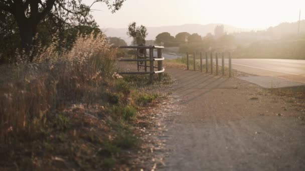 Hombre en bicicleta negra a lo largo de un camino rural al atardecer. Hermoso campo en luz dorada. Campos dorados con cercas de madera y colinas verdes. Estilo de vida saludable en la naturaleza. — Vídeo de stock