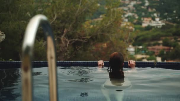 Fille aux cheveux longs se détendre dans la piscine et admirer la belle vue. Une jeune femme aux cheveux mouillés passe une journée d'été dans l'eau bleue fraîche. Vue sur les arbres verts et les montagnes. Refroidissement estival. — Video