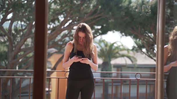Een langharige vrouw met zwarte legging en een T-shirt staat op het balkon met een telefoon. Het meisje sms 't een bericht aan de telefoon. Mooie bomen achter haar. Communicatie. Het internet. Mobiele telefoon. — Stockvideo